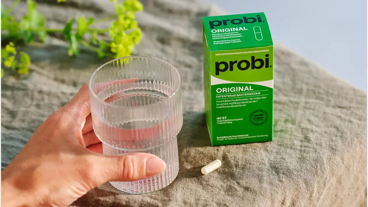 En hand håller ett glas vatten bredvid en ask med Probi Original. Förpackningen förklarar vad innehåller probiotika: levande mjölksyrabakterier.
