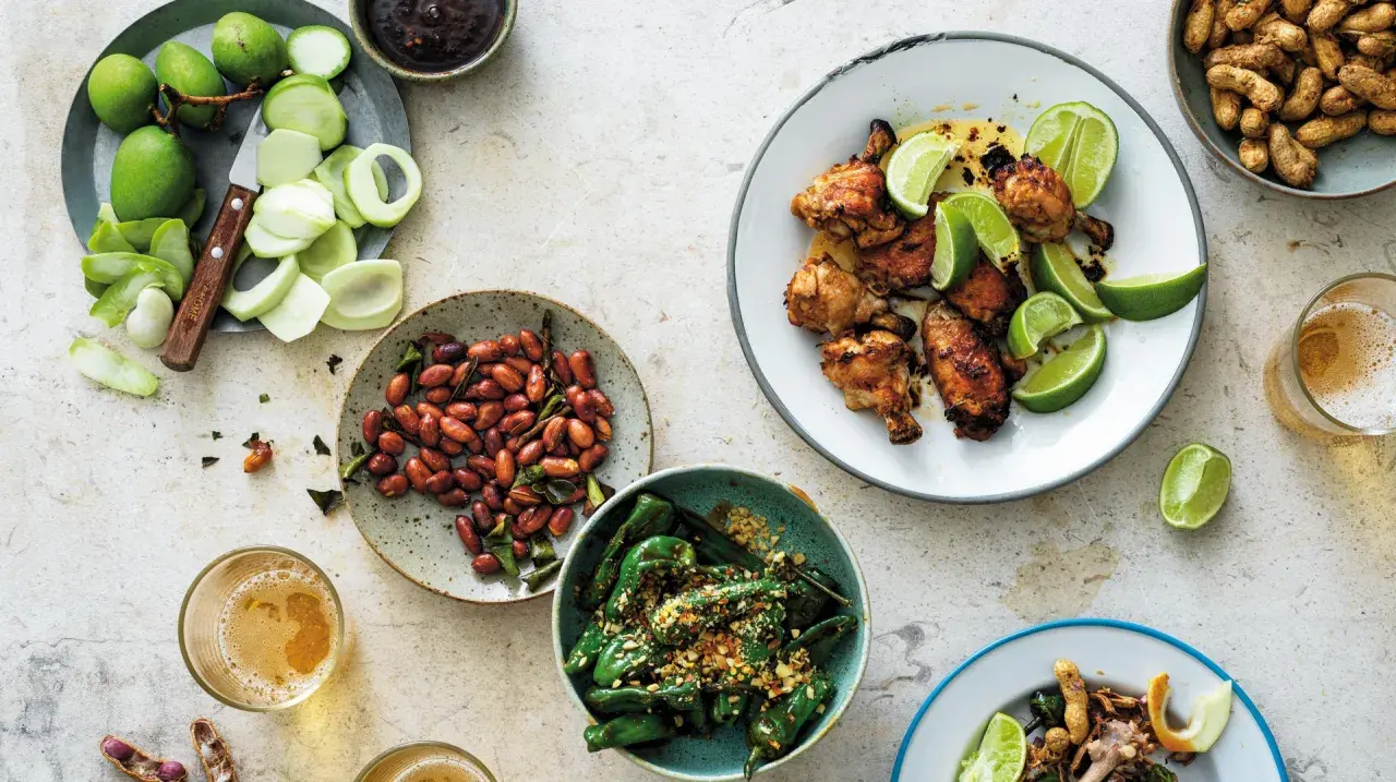 En härlig dukning med olika rätter av thai mat: kycklingvingar, friterade gröna paprikor, jordnötter och omogna mangos.