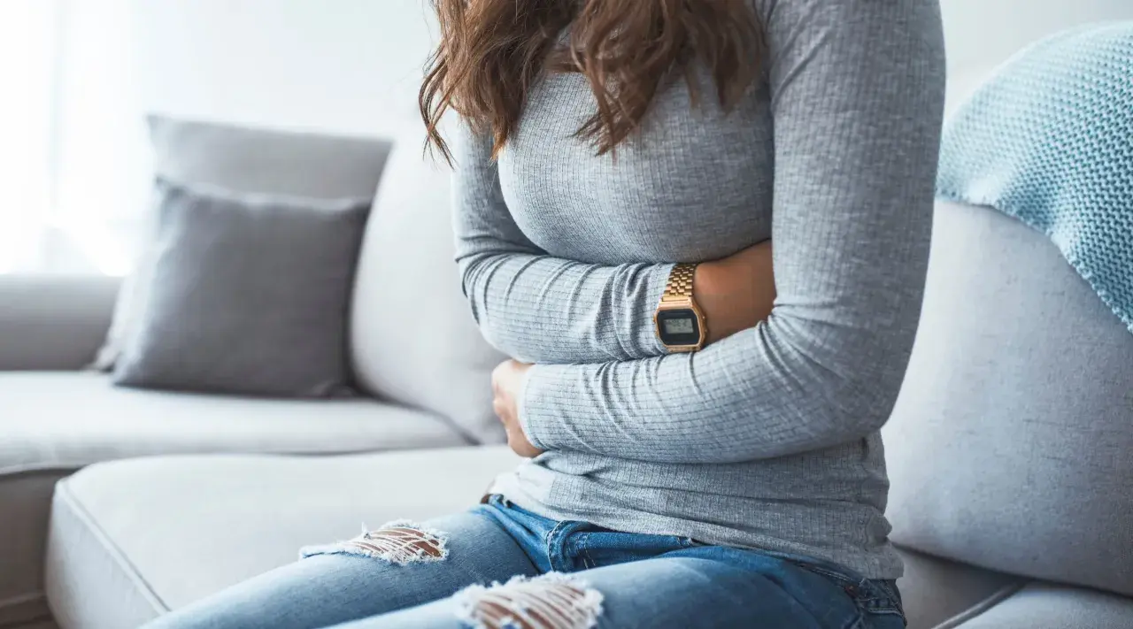 Kvinna med händerna på magen, upplever symtom på IBS. Hon sitter på en soffa med en filt i bakgrunden.