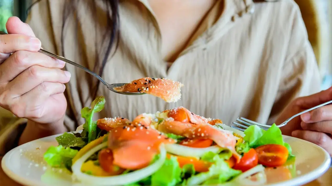 En person äter en fräsch sallad med lax, tomater och lök. En sked med lax, strödd med sesamfrön, lyfts mot munnen. Detta är ett exempel på q10 mat.