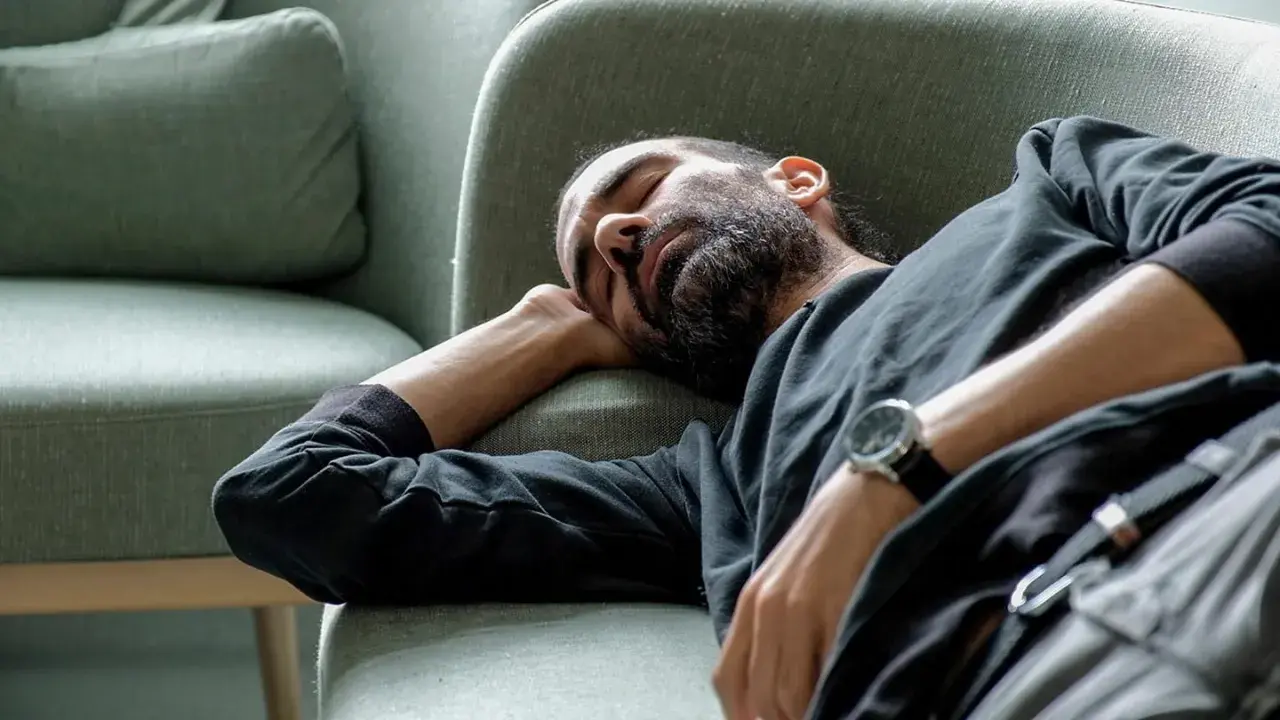 Man med skägg sover på en soffa, med armen under huvudet. Han verkar utmattad, kanske av illamående som kommer och går.