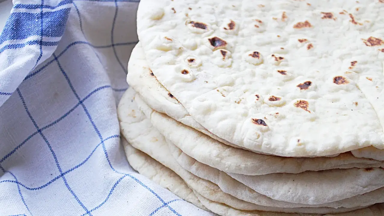 En hög med nybakade tortillas, perfekta för ditt nästa tortilla recept. En blårutig kökshandduk ligger bredvid.