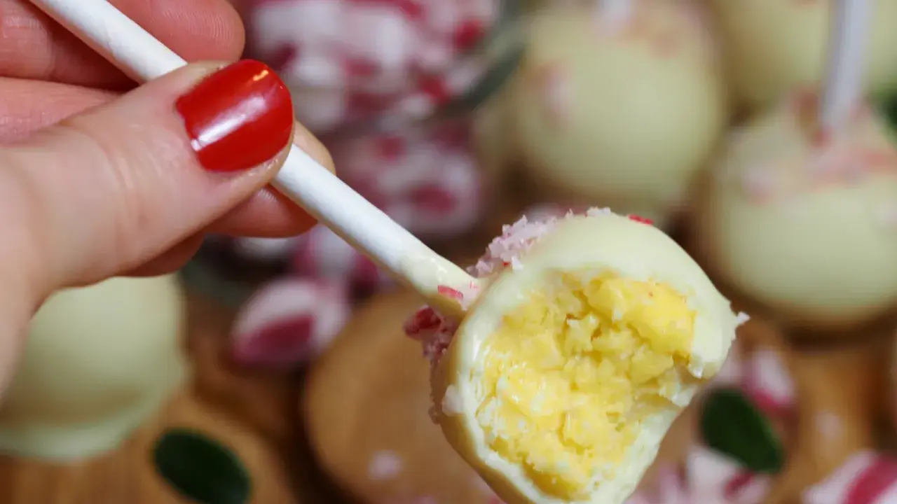 En hand med rött nagellack håller en cake pop. Den är doppad i vit choklad och strössel.