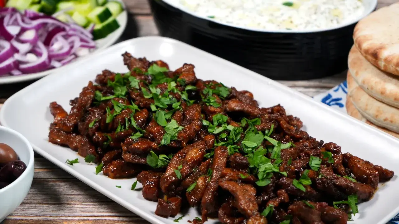 En tallrik med saftigt gyroskött, toppat med färsk persilja. Bredvid syns pitabröd, tzatziki, oliver, gurka och rödlök. Ett perfekt gyros recept!