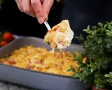 En sked lyfter upp en portion gyllene makaronilåda med korv ur en ugnsform.