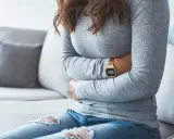 Kvinna med händerna på magen, upplever symtom på IBS. Hon sitter på en soffa med en filt i bakgrunden.