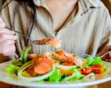 En person äter en fräsch sallad med lax, tomater och lök. En sked med lax, strödd med sesamfrön, lyfts mot munnen. Detta är ett exempel på q10 mat.