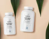 Två burkar med Holistic magsyra-balans och måltids-enzym, omgivna av gröna blad.