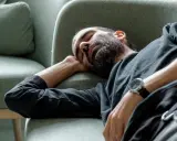 Man med skägg sover på en soffa, med armen under huvudet. Han verkar utmattad, kanske av illamående som kommer och går.