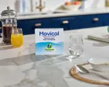 En låda Movicol med citron- och limsmak står på en köksbänk. Bredvid finns ett glas vatten, juice och en presskanna kaffe. Tänk på moxalole verkningstid för effektiv lindring.