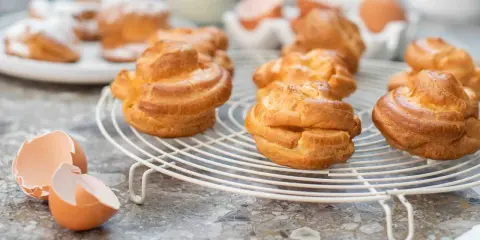 Gyllene petit choux på ett vitt galler, med äggskal och ägg i bakgrunden.