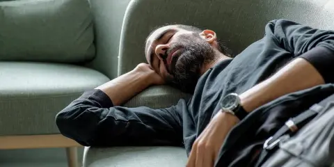 Man med skägg sover på en soffa, med armen under huvudet. Han verkar utmattad, kanske av illamående som kommer och går.