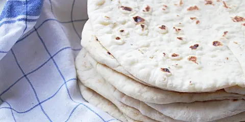 En hög med nybakade tortillas, perfekta för ditt nästa tortilla recept. En blårutig kökshandduk ligger bredvid.