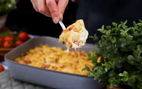 En sked lyfter upp en portion gyllene makaronilåda med korv ur en ugnsform.