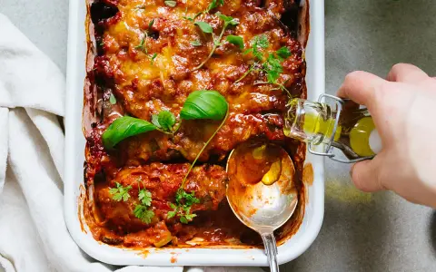 En ugnsbakad auberginegratäng med tomatsås och ost, toppad med färska örter. En sked och en flaska olivolja antyder ett läckert aubergine recept.