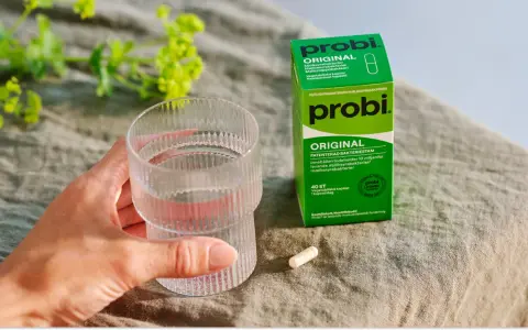 En hand håller ett glas vatten bredvid en förpackning med probi. En kapsel ligger på bordet, redo att tas för att undvika probi mage biverkningar.
