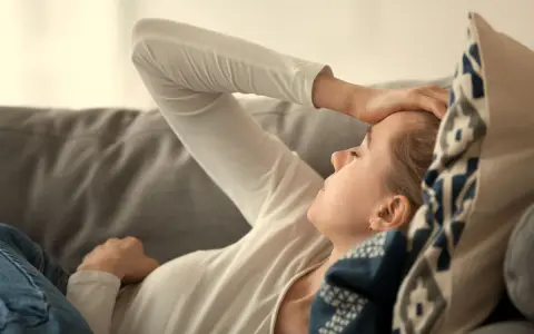 Kvinna med handen på pannan, kanske ett tecken på b12-brist symtom som trötthet eller huvudvärk. Hon vilar på en soffa.