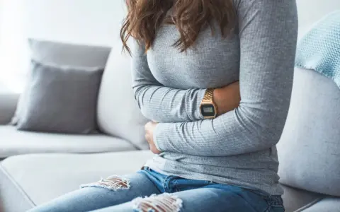 Kvinna med händerna på magen, upplever symtom på IBS. Hon sitter på en soffa med en filt i bakgrunden.