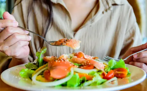 En person äter en fräsch sallad med lax, tomater och lök. En sked med lax, strödd med sesamfrön, lyfts mot munnen. Detta är ett exempel på q10 mat.