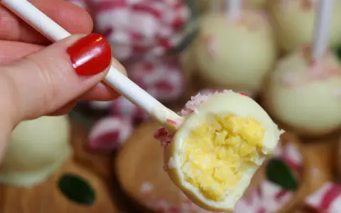 En hand med rött nagellack håller en cake pop. Den är doppad i vit choklad och strössel.