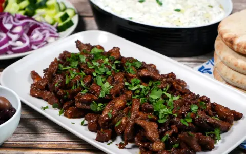 En tallrik med saftigt gyroskött, toppat med färsk persilja. Bredvid syns pitabröd, tzatziki, oliver, gurka och rödlök. Ett perfekt gyros recept!