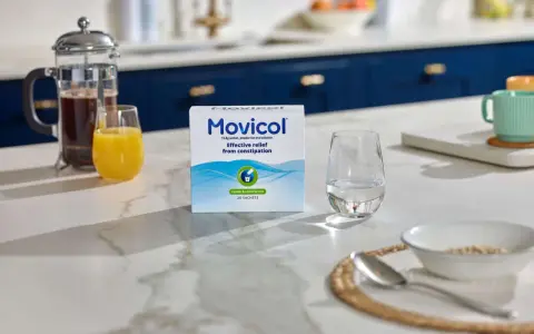 En låda Movicol med citron- och limsmak står på en köksbänk. Bredvid finns ett glas vatten, juice och en presskanna kaffe. Tänk på moxalole verkningstid för effektiv lindring.