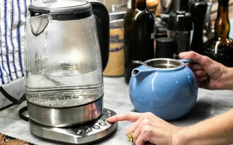 En person trycker på knappen för att värma vatten i en vattenkokare, redo att dricka varmt vatten på morgonen. En blå tekanna står bredvid.