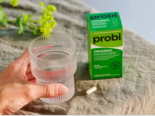 En hand håller ett glas vatten bredvid en ask med Probi Original. Förpackningen förklarar vad innehåller probiotika: levande mjölksyrabakterier.