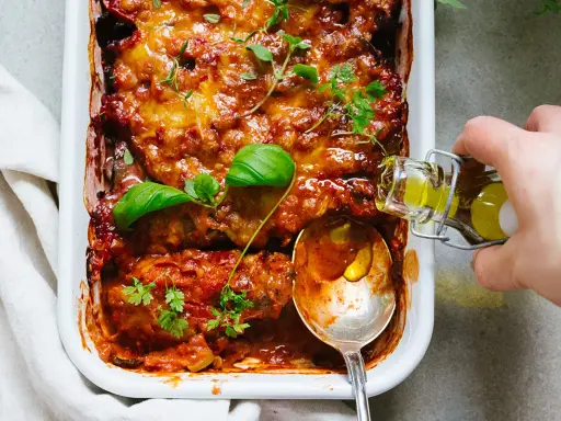 En ugnsbakad auberginegratäng med tomatsås och ost, toppad med färska örter. En sked och en flaska olivolja antyder ett läckert aubergine recept.
