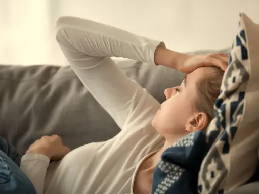 Kvinna med handen på pannan, kanske ett tecken på b12-brist symtom som trötthet eller huvudvärk. Hon vilar på en soffa.