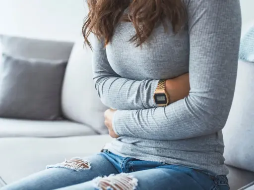 Kvinna med händerna på magen, upplever symtom på IBS. Hon sitter på en soffa med en filt i bakgrunden.