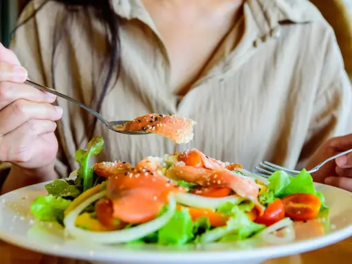 En person äter en fräsch sallad med lax, tomater och lök. En sked med lax, strödd med sesamfrön, lyfts mot munnen. Detta är ett exempel på q10 mat.
