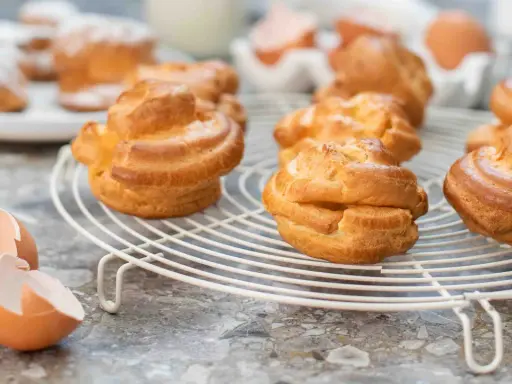 Gyllene petit choux på ett vitt galler, med äggskal och ägg i bakgrunden.