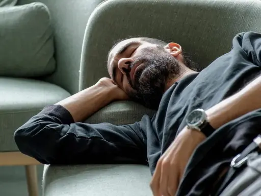 Man med skägg sover på en soffa, med armen under huvudet. Han verkar utmattad, kanske av illamående som kommer och går.