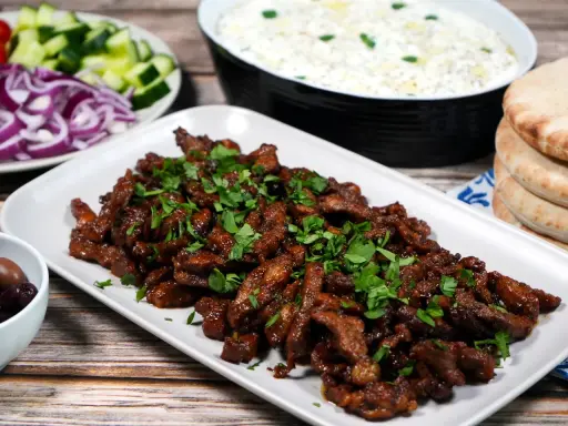 En tallrik med saftigt gyroskött, toppat med färsk persilja. Bredvid syns pitabröd, tzatziki, oliver, gurka och rödlök. Ett perfekt gyros recept!