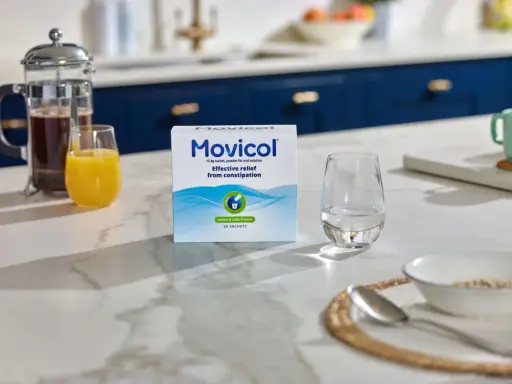 En låda Movicol med citron- och limsmak står på en köksbänk. Bredvid finns ett glas vatten, juice och en presskanna kaffe. Tänk på moxalole verkningstid för effektiv lindring.