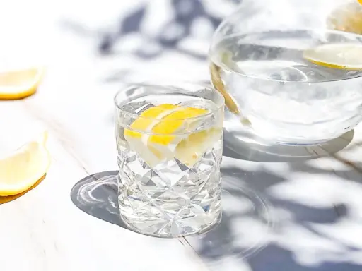 Ett glas med vatten med elektrolyter och citronskivor, bredvid en karaff med samma dryck. Solens strålar skapar vackra skuggor.