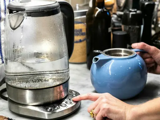 En person trycker på knappen för att värma vatten i en vattenkokare, redo att dricka varmt vatten på morgonen. En blå tekanna står bredvid.