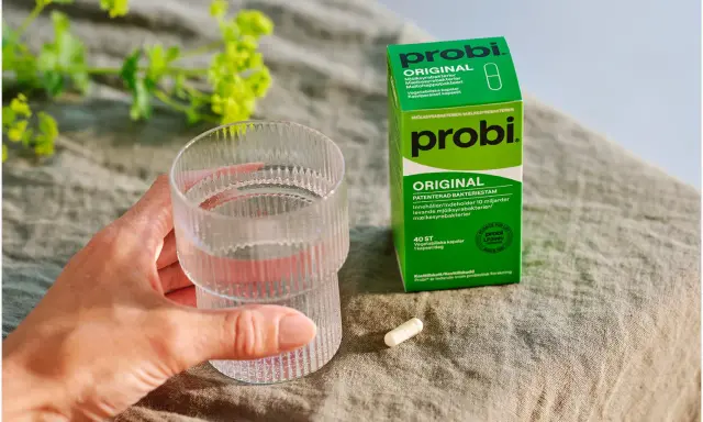 En hand håller ett glas vatten bredvid en ask med Probi Original. Förpackningen förklarar vad innehåller probiotika: levande mjölksyrabakterier.