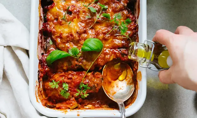 En ugnsbakad auberginegratäng med tomatsås och ost, toppad med färska örter. En sked och en flaska olivolja antyder ett läckert aubergine recept.