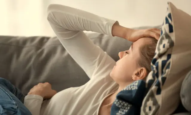Kvinna med handen på pannan, kanske ett tecken på b12-brist symtom som trötthet eller huvudvärk. Hon vilar på en soffa.