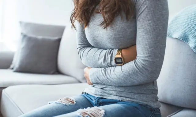 Kvinna med händerna på magen, upplever symtom på IBS. Hon sitter på en soffa med en filt i bakgrunden.