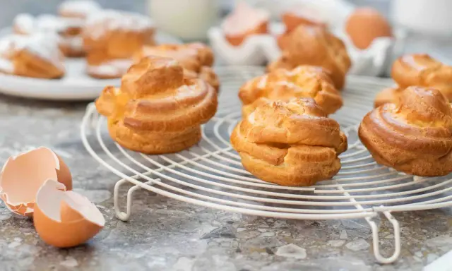 Gyllene petit choux på ett vitt galler, med äggskal och ägg i bakgrunden.