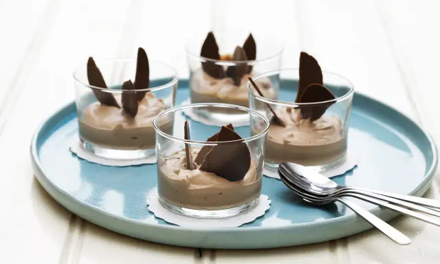 Fyra glas med enkel chokladmousse, dekorerade med chokladflarn, står på en turkos bricka. Bredvid ligger en hög med skedar.