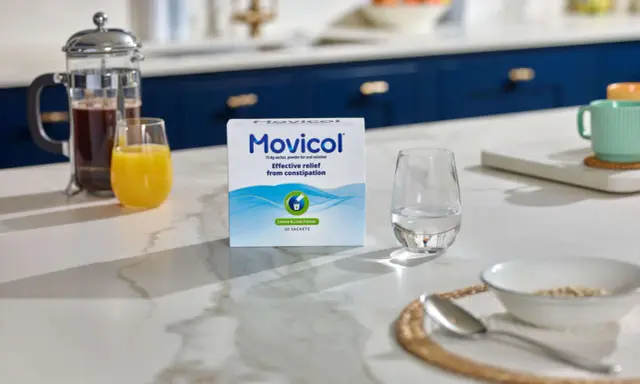 En låda Movicol med citron- och limsmak står på en köksbänk. Bredvid finns ett glas vatten, juice och en presskanna kaffe. Tänk på moxalole verkningstid för effektiv lindring.