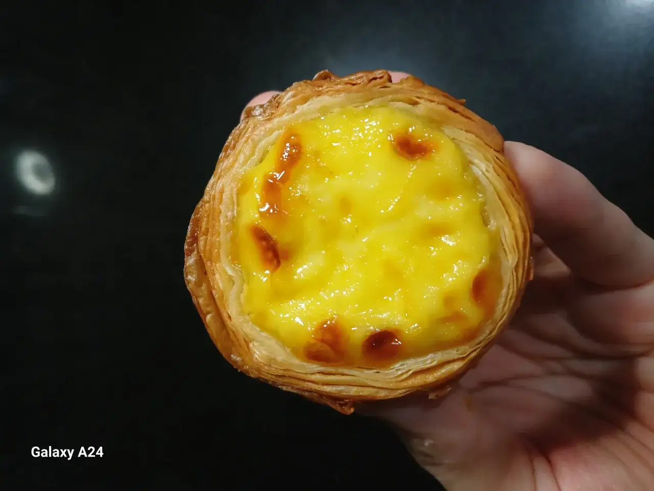 En hand h&aring;ller en gyllene, krispig pastel de nata med kr&auml;mig vaniljkr&auml;m och karamelliserade toppar.