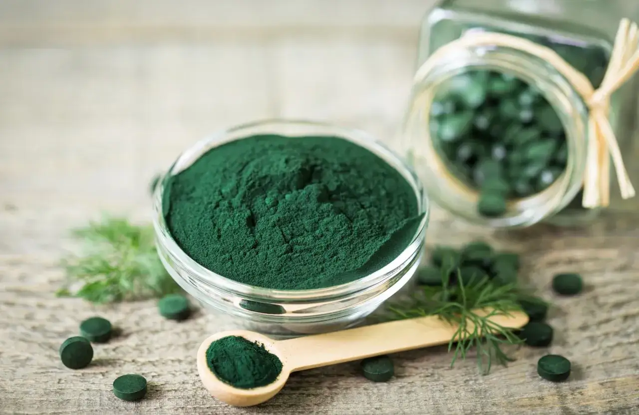 Gr&ouml;n spirulinapulver i sk&aring;l och kapslar i burk. T&auml;nk p&aring; eventuella spirulina biverkningar vid anv&auml;ndning.