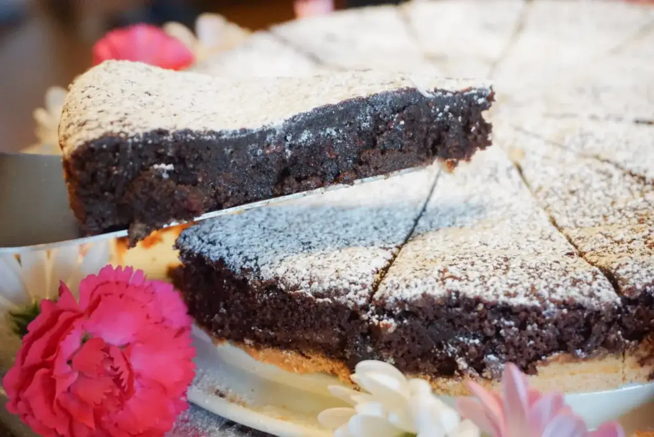 En bit av en h&auml;rlig glutenfri kladdkaka lyfts upp. Den &auml;r pudrad med florsocker och dekorerad med rosa och vita blommor.