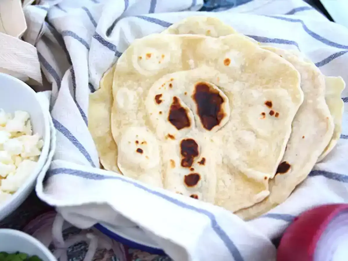 F&auml;rska, hembakade tortillas p&aring; en randig handduk, redo f&ouml;r ditt b&auml;sta tortilla recept. Bredvid syns sk&aring;lar med ost och l&ouml;k.
