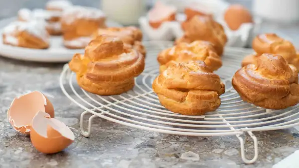 Gyllene petit choux på ett vitt galler, med äggskal och ägg i bakgrunden.